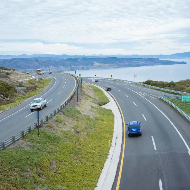 Autopista costera con varios vehículos en movimiento, bordeada de vegetación y con vista al mar y montañas en el fondo. Señales indican direcciones a Playa Saldamando y Ensenada.